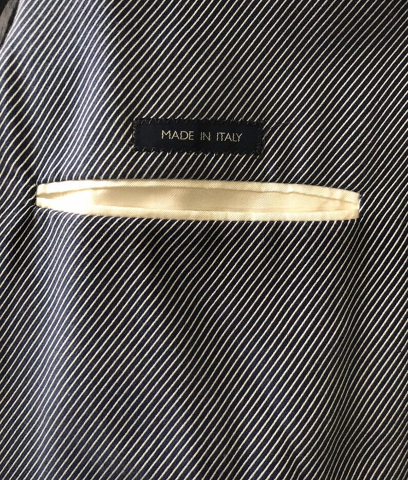 May include: Close-up of the interior lining of a dark navy blue blazer with white pinstripes. A small, rectangular label with the text "MADE IN ITALY" is sewn into the lining. A cream-colored pocket is visible.