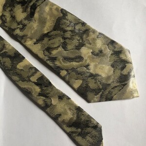 Puede incluir: Una corbata con un patr&oacute;n abstracto y moteado en tonos verde oliva, marr&oacute;n oscuro y crema. La tela tiene una textura tejida sutil con un ligero brillo, mostrando una punta afilada y una forma larga y c&oacute;nica con un extremo estrecho a juego.