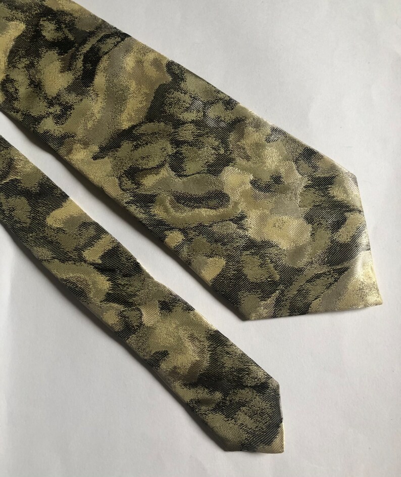 Puede incluir: Una corbata con un patr&oacute;n abstracto y moteado en tonos verde oliva, marr&oacute;n oscuro y crema. La tela tiene una textura tejida sutil con un ligero brillo, mostrando una punta afilada y una forma larga y c&oacute;nica con un extremo estrecho a juego.