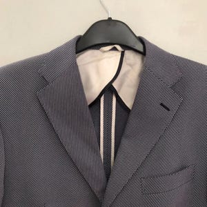 May include: A blue and white striped blazer hanging on a black hanger. The blazer has a notched lapel, a chest pocket, and a two-button closure. The interior lining is white with black trim.