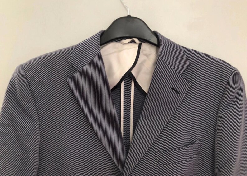 May include: A blue and white striped blazer hanging on a black hanger. The blazer has a notched lapel, a chest pocket, and a two-button closure. The interior lining is white with black trim.
