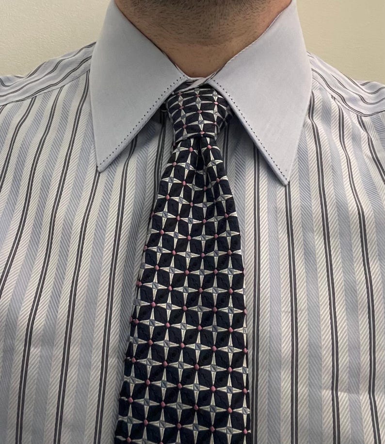 Pu&ograve; includere: Una camicia elegante a righe azzurre e bianche con un colletto azzurro a tinta unita caratterizzato da cuciture decorative lungo i bordi. La camicia &egrave; abbinata a una cravatta blu navy scuro con un motivo geometrico a stella in bianco e rosa.