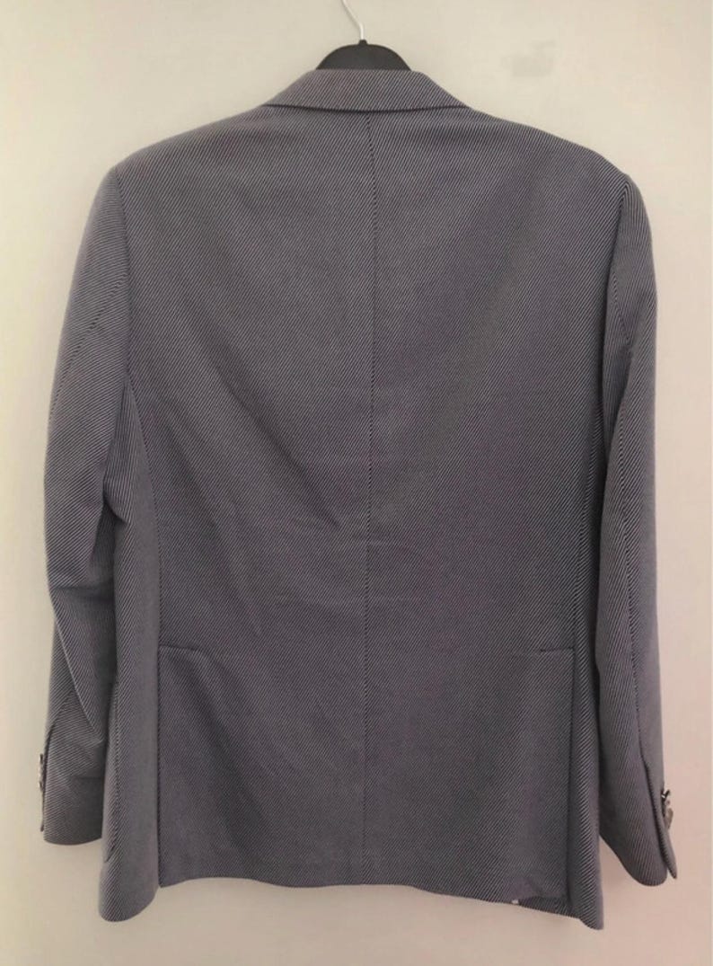 May include: A gray blazer with a subtle, textured pattern. The jacket is hung on a black hanger and features a classic two-button closure, a notched lapel, and a single vent in the back. The fabric appears to be a lightweight material.
