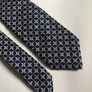 Pu&ograve; includere: Una cravatta con sfondo blu navy scuro e un motivo geometrico ripetitivo di stelle bianche a quattro punte e piccoli punti rosa. Il tessuto presenta una trama testurizzata e la cravatta &egrave; mostrata con la pala larga e l'estremit&agrave; stretta visibili.