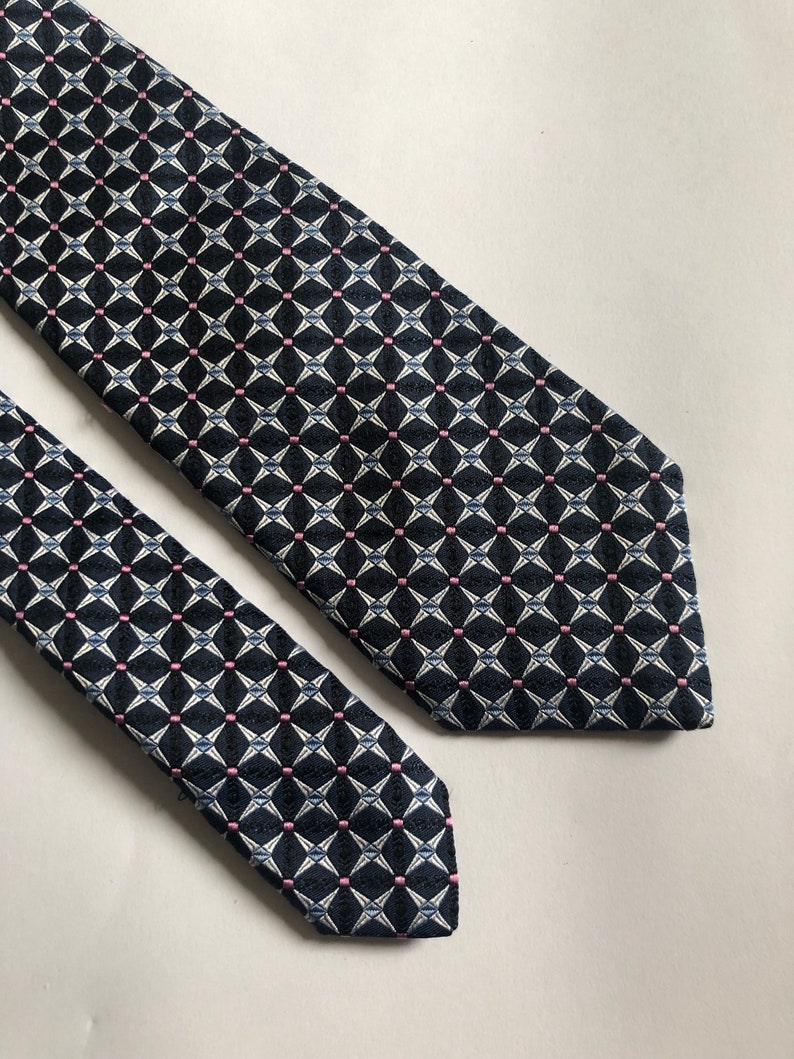 Pu&ograve; includere: Una cravatta con sfondo blu navy scuro e un motivo geometrico ripetitivo di stelle bianche a quattro punte e piccoli punti rosa. Il tessuto presenta una trama testurizzata e la cravatta &egrave; mostrata con la pala larga e l'estremit&agrave; stretta visibili.