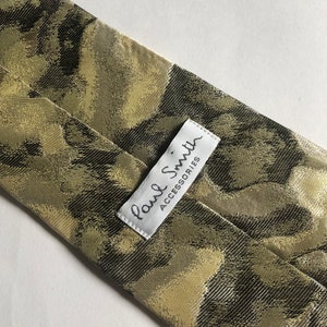 Puede incluir: Una corbata con un patr&oacute;n abstracto similar al camuflaje en tonos verde oliva, marr&oacute;n oscuro y bronceado. Una etiqueta de tela rectangular blanca est&aacute; cosida en la parte posterior, mostrando el texto Paul Smith ACCESSORIES en letras negras.