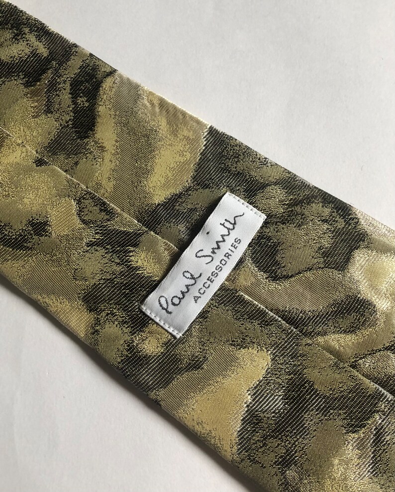 Puede incluir: Una corbata con un patr&oacute;n abstracto similar al camuflaje en tonos verde oliva, marr&oacute;n oscuro y bronceado. Una etiqueta de tela rectangular blanca est&aacute; cosida en la parte posterior, mostrando el texto Paul Smith ACCESSORIES en letras negras.