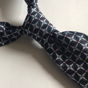 Pu&ograve; includere: Una cravatta con una base blu navy scuro e un motivo geometrico ripetitivo di forme a stella bianche e piccoli punti rosa. Il tessuto ha un aspetto strutturato con un nodo ben definito legato nella parte superiore, mostrando la pala.