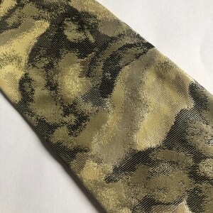 Puede incluir: Una corbata de tela tejida con un patr&oacute;n abstracto y texturizado en tonos verde oliva, marr&oacute;n oscuro y oro met&aacute;lico. El material presenta un tejido de sarga diagonal en toda la superficie, creando un brillo sutil sobre la corbata.