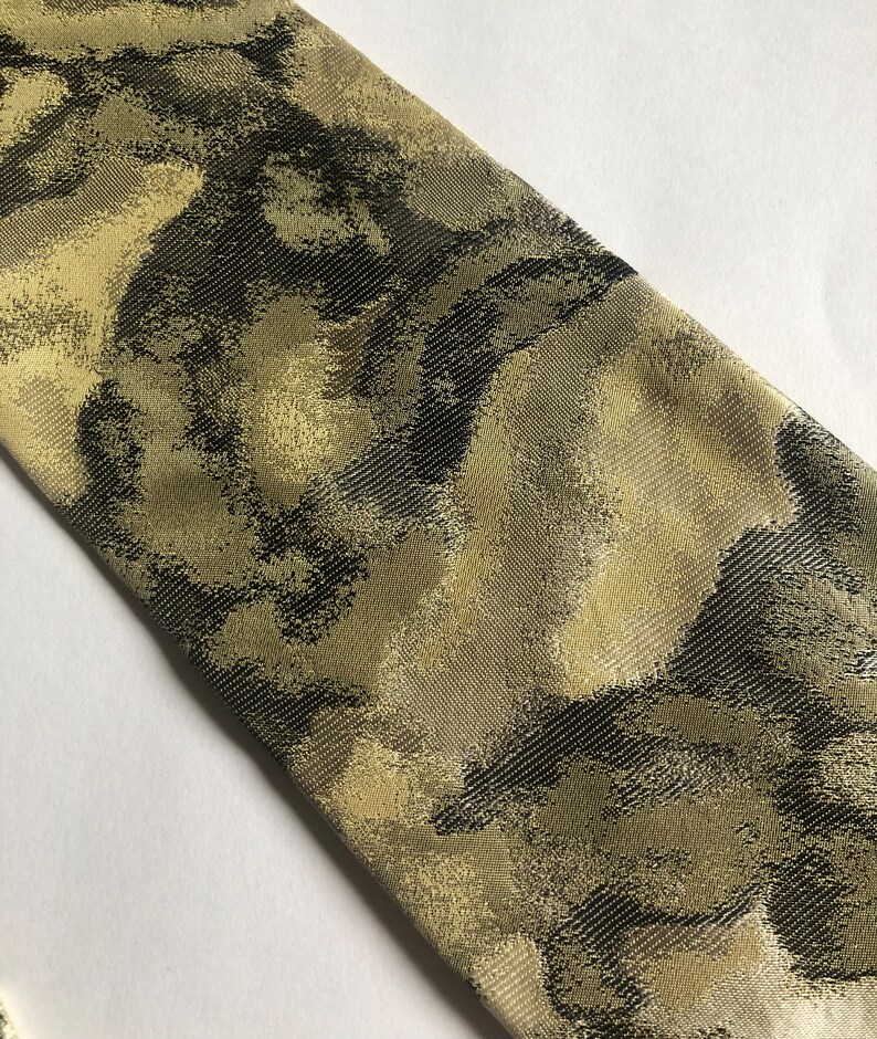 Puede incluir: Una corbata de tela tejida con un patr&oacute;n abstracto y texturizado en tonos verde oliva, marr&oacute;n oscuro y oro met&aacute;lico. El material presenta un tejido de sarga diagonal en toda la superficie, creando un brillo sutil sobre la corbata.
