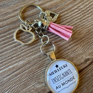May include: A keyring featuring a round pendant with the text MEILLEURE ENSEIGNANTE AU MONDE and a star icon, accompanied by a silver-toned apple charm, a small heart charm, and a pink suede tassel attached to a circular metal ring.