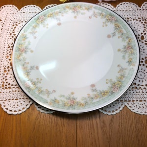 May include: A white ceramic dinner plate with a green and yellow floral pattern around the rim. The plate has a silver trim.