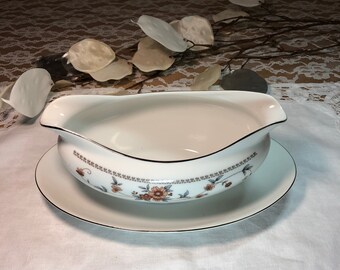 May include: A white porcelain gravy boat with a floral design and a gold trim. The boat is sitting on a matching saucer.