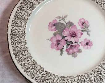 May include: A white ceramic plate with a gold rim and a floral design featuring pink flowers and green leaves. The plate has a delicate, intricate gold pattern around the edge.