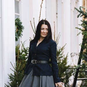 May include: A woman wearing a dark blue button-up shirt with a black belt and a long, gray skirt. She is standing in front of a white building with greenery.