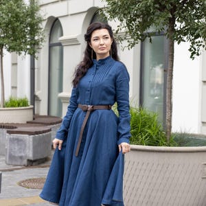 May include: A long-sleeved, blue dress with a high collar and button-down front. The dress is cinched at the waist with a brown belt and has a flowing skirt. The person is wearing black boots. The dress appears to be made of linen or a similar fabric.