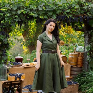 May include: A woman in a green dress with a brown belt stands in a vineyard setting. The dress has a button-down front and short sleeves. Grapes, wine, and wooden crates are visible, suggesting a wine-tasting theme. The scene is outdoors with lush greenery.