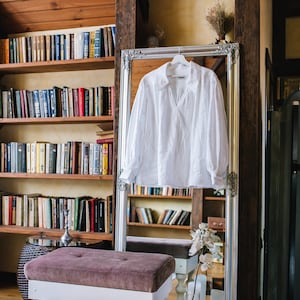 May include: A white long-sleeved shirt with a V-neck hangs in front of a mirror with an ornate silver frame. A tufted bench with white legs is in front of the mirror. Bookshelves filled with books are in the background.