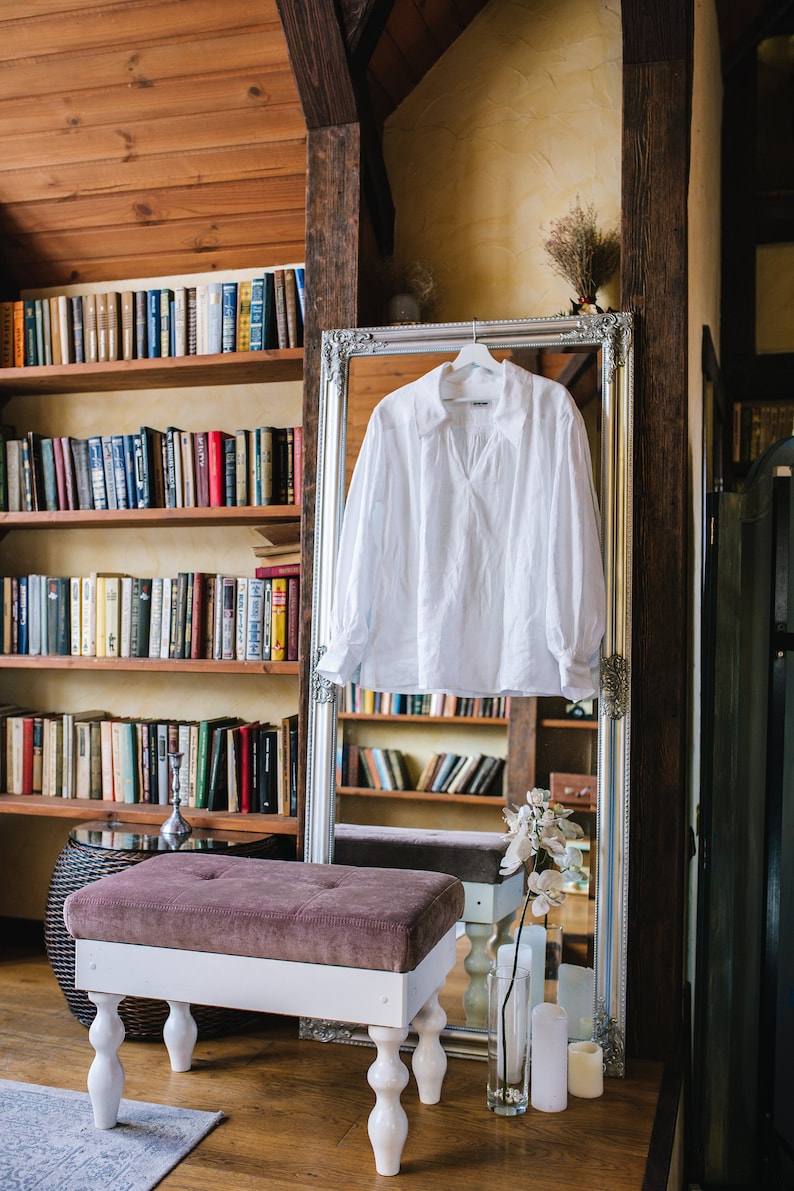 May include: A white long-sleeved shirt with a V-neck hangs in front of a mirror with an ornate silver frame. A tufted bench with white legs is in front of the mirror. Bookshelves filled with books are in the background.
