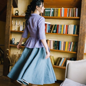 Op de afbeelding: Een vrouw draagt een lichtpaarse linnen blouse met een gerimpelde zoom en een lichtblauwe linnen rok. Ze staat voor een boekenkast vol boeken.