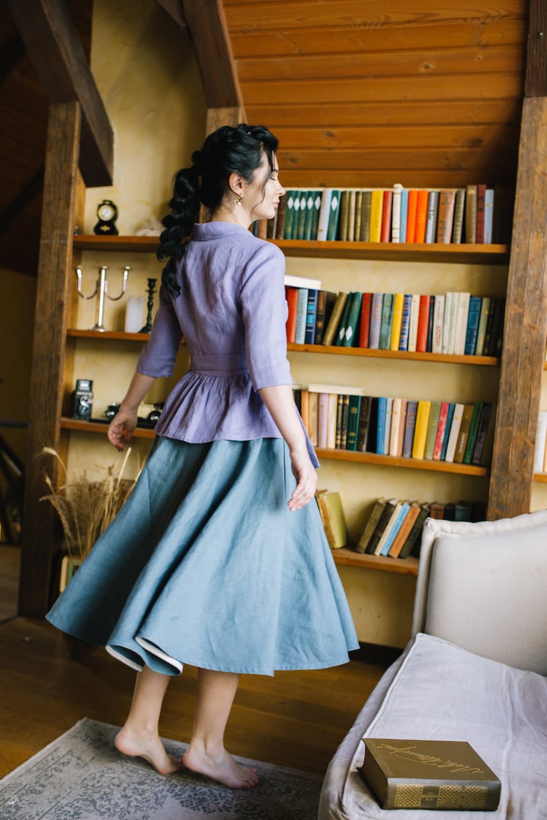 Op de afbeelding: Een vrouw draagt een lichtpaarse linnen blouse met een gerimpelde zoom en een lichtblauwe linnen rok. Ze staat voor een boekenkast vol boeken.
