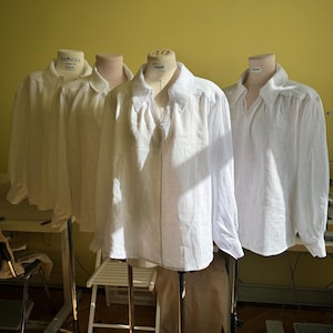 May include: Three white linen shirts displayed on dressmaker's dummies. The shirts feature long sleeves and a classic collar. The dummies are on black metal stands, against a yellow wall. Natural light illuminates the scene.