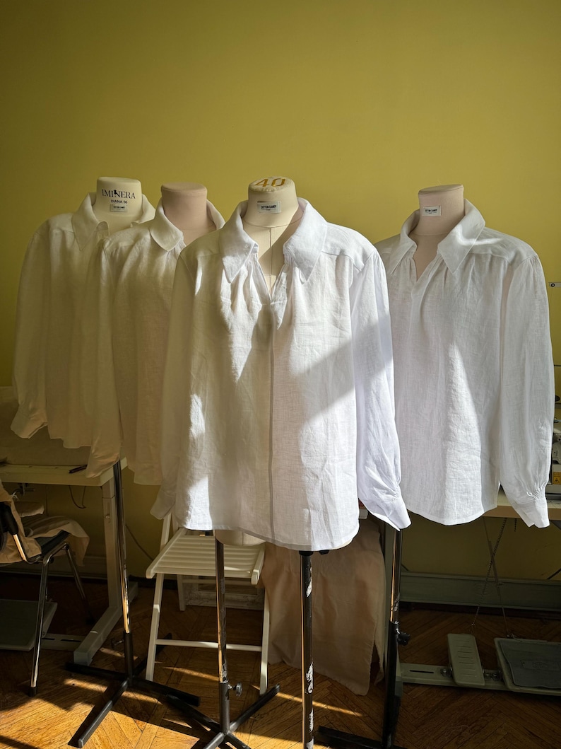 May include: Three white linen shirts displayed on dressmaker's dummies. The shirts feature long sleeves and a classic collar. The dummies are on black metal stands, against a yellow wall. Natural light illuminates the scene.