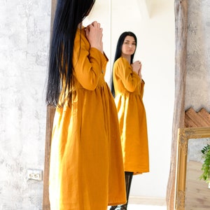 May include: A woman in a mustard yellow linen dress stands in front of a mirror, looking at her reflection. She is wearing black boots and has long black hair.