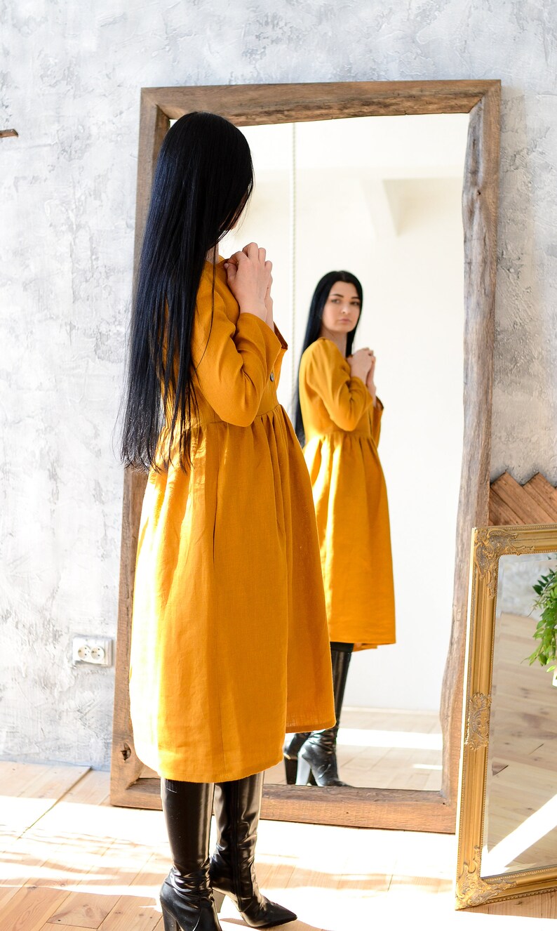May include: A woman in a mustard yellow linen dress stands in front of a mirror, looking at her reflection. She is wearing black boots and has long black hair.