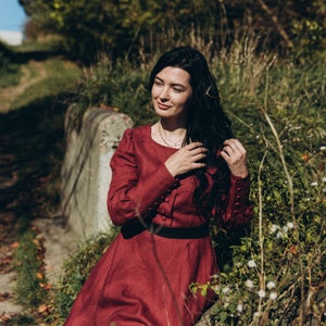 May include: A woman in a red dress sits on a stone wall in a field of white flowers. She is wearing a necklace and has long black hair.