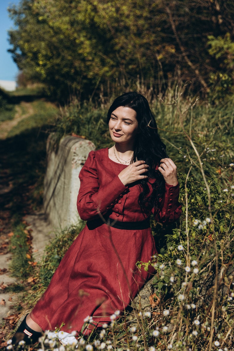 May include: A woman in a red dress sits on a stone wall in a field of white flowers. She is wearing a necklace and has long black hair.
