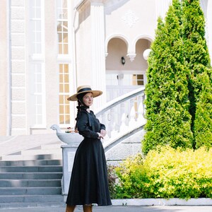 May include: A woman wearing a black dress with a wide skirt and a straw hat stands in front of a white building with a staircase. She is wearing brown boots and has her hair pulled back.