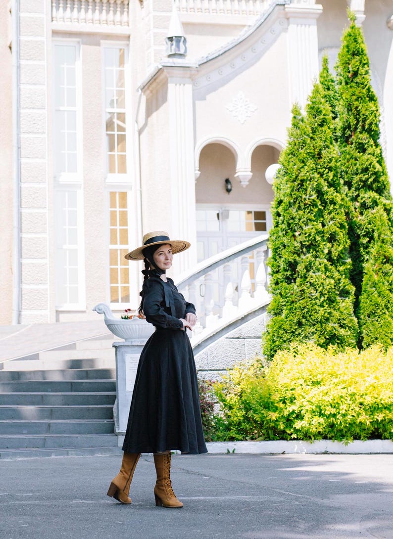 May include: A woman wearing a black dress with a wide skirt and a straw hat stands in front of a white building with a staircase. She is wearing brown boots and has her hair pulled back.