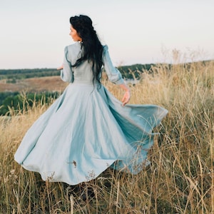 Puede incluir: Una mujer con un vestido azul claro gira en un campo de hierba alta. El vestido es fluido y tiene una falda amplia. El cabello de la mujer es largo y oscuro.