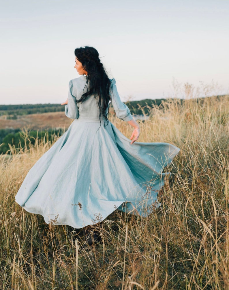 Puede incluir: Una mujer con un vestido azul claro gira en un campo de hierba alta. El vestido es fluido y tiene una falda amplia. El cabello de la mujer es largo y oscuro.