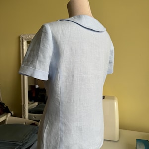 Puede incluir: Una blusa azul claro de manga corta con cuello redondo se exhibe en un maniqu&iacute;. La blusa est&aacute; hecha de una tela texturizada y las mangas tienen un pu&ntilde;o doblado. La parte posterior de la blusa es visible, mostrando las costuras a medida.