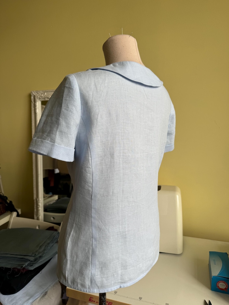 Puede incluir: Una blusa azul claro de manga corta con cuello redondo se exhibe en un maniqu&iacute;. La blusa est&aacute; hecha de una tela texturizada y las mangas tienen un pu&ntilde;o doblado. La parte posterior de la blusa es visible, mostrando las costuras a medida.