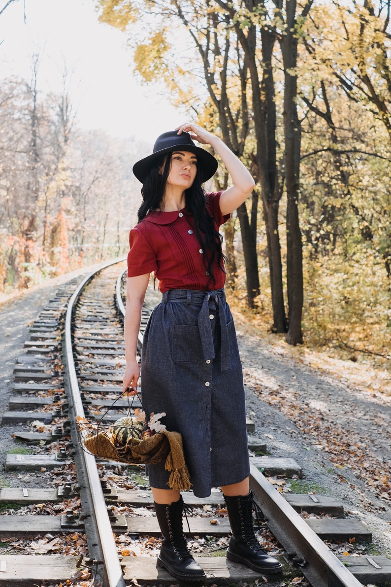 Puede incluir: Una persona con una blusa burdeos de manga corta y una falda vaquera con cintur&oacute;n, de pie sobre v&iacute;as de tren. Sostiene una cesta con calabazas y una bufanda. Lleva un sombrero y botas negras. El fondo muestra &aacute;rboles con hojas amarillas.