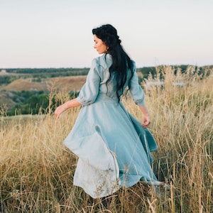 Puede incluir: Una mujer con un vestido de lino azul claro camina por un campo de hierba alta.