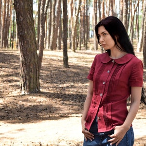 Puede incluir: Una blusa burdeos de manga corta con cuello redondo y cierre de botones. Pliegues verticales adornan el centro de la blusa. La blusa se combina con vaqueros azules. El fondo es un bosque con &aacute;rboles altos.