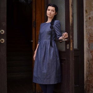 Puede incluir: Una mujer con un vestido de mezclilla azul se para en una puerta. Lleva medias negras y botas negras con cordones. La puerta es de madera oscura y tiene un panel de vidrio.