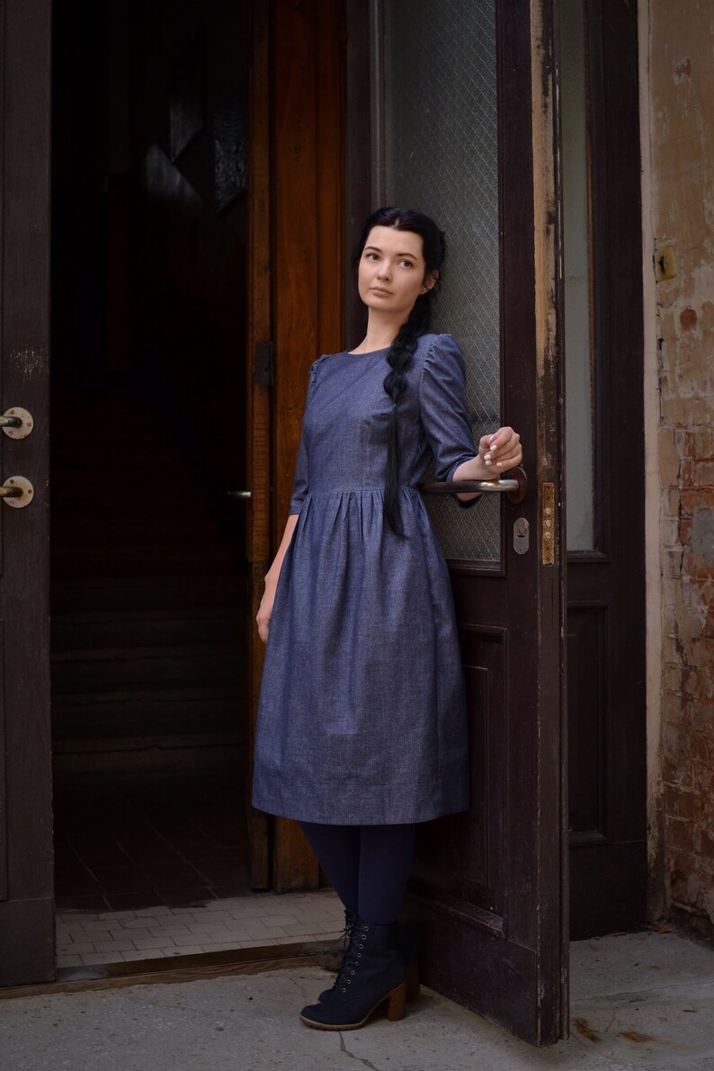 Puede incluir: Una mujer con un vestido de mezclilla azul se para en una puerta. Lleva medias negras y botas negras con cordones. La puerta es de madera oscura y tiene un panel de vidrio.