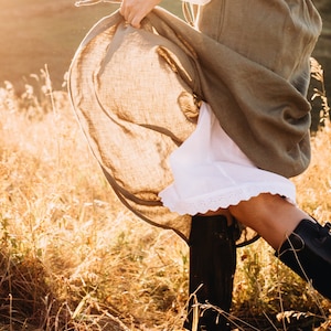 May include: A woman wearing a long, brown linen dress with a white underskirt and lace trim. She is standing in a field of tall grass with the sun shining on her.