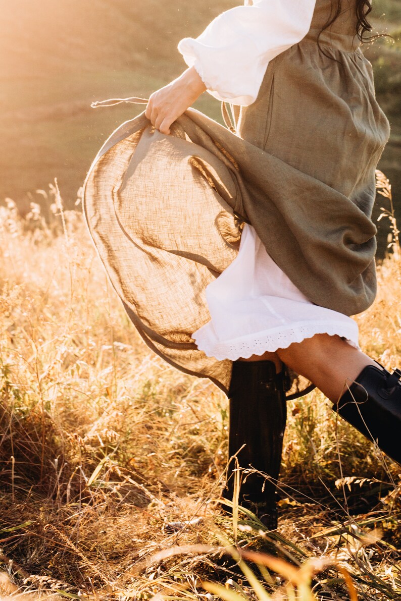 May include: A woman wearing a long, brown linen dress with a white underskirt and lace trim. She is standing in a field of tall grass with the sun shining on her.