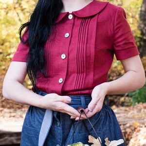Puede incluir: Una blusa burdeos de manga corta con cuello Peter Pan y cierre de botones. La blusa tiene pliegues verticales en el centro. Tambi&eacute;n se ve una falda vaquera y una cesta de alambre llena de calabazas y manzanas.