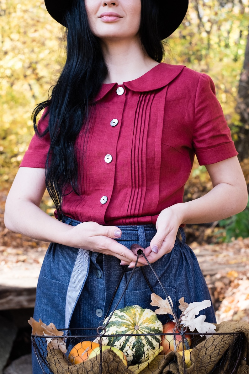 Puede incluir: Una blusa burdeos de manga corta con cuello Peter Pan y cierre de botones. La blusa tiene pliegues verticales en el centro. Tambi&eacute;n se ve una falda vaquera y una cesta de alambre llena de calabazas y manzanas.