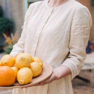 Pu&ograve; includere: Una donna con un abito di lino beige chiaro tiene un vassoio di legno pieno di limoni e arance. I limoni sono gialli e le arance sono arancioni. La donna sorride e guarda la telecamera.