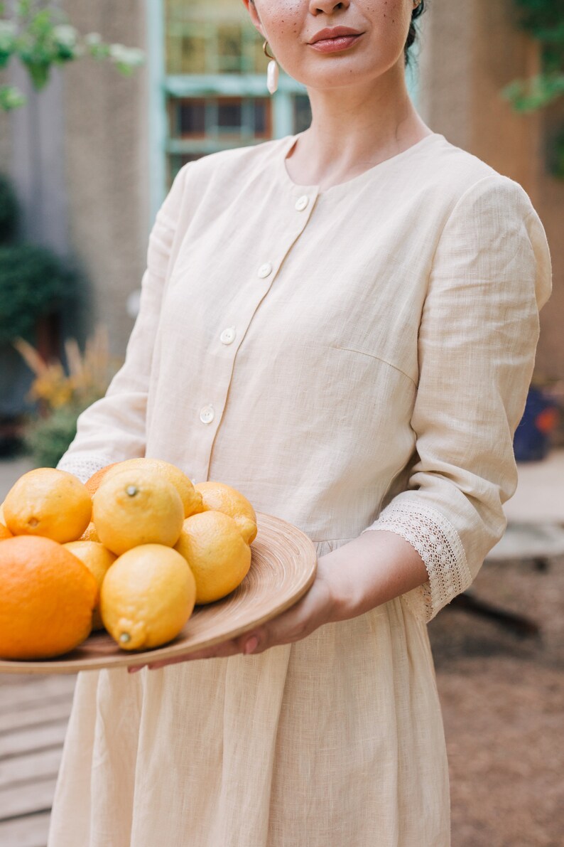 Pu&ograve; includere: Una donna con un abito di lino beige chiaro tiene un vassoio di legno pieno di limoni e arance. I limoni sono gialli e le arance sono arancioni. La donna sorride e guarda la telecamera.