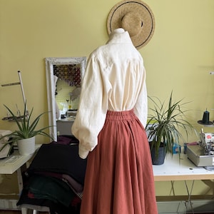 May include: A cream linen blouse with long, puffed sleeves and a high collar is styled with a rust-coloured, full-length skirt. The skirt has a gathered waist. A straw hat hangs on the wall behind the outfit.