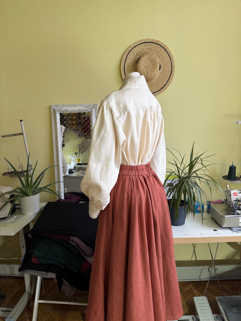 May include: A cream linen blouse with long, puffed sleeves and a high collar is styled with a rust-coloured, full-length skirt. The skirt has a gathered waist. A straw hat hangs on the wall behind the outfit.